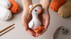 Ganso amigurumi de crochê branco com detalhes em laranja, em pé sobre uma mesa, acessório de decoração artesanal.