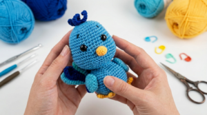 Pavão Pablo Amigurumi com plumagem azul e turquesa feito em crochê, usado como peça decorativa charmosa em estante.