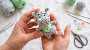 Mini hipopótamo de crochê cinza suave feito à mão, posicionado em ambiente decorativo para lembrancinha ou chaveiro artesanal.